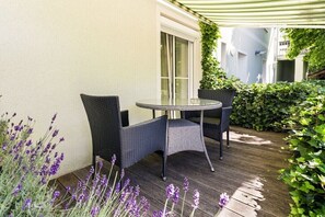 Eine Terrasse im Freien mit Korbmöbeln, einem Glastisch und Blick auf ein Gebäude mit weißer Fassade und einem grünen Markisenvorbau.