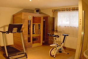 Ein Zimmer mit Laufband, Ergometer, Holzschrank und einem Fenster mit Vorhängen.