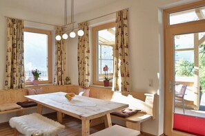 Ein Zimmer mit Holztisch, Bänken und großen Fenstern mit Vorhängen.