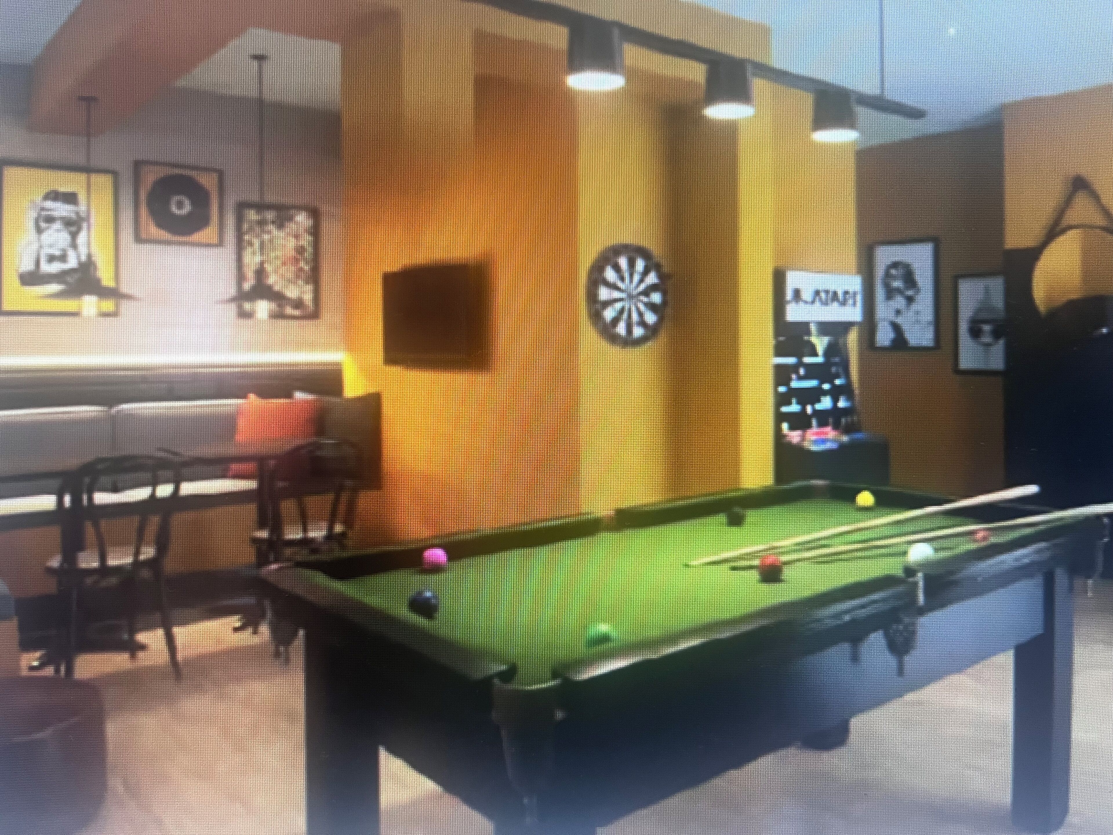 Mesa de bilhar com dardos e jukebox num espaço com sofá e cadeiras.