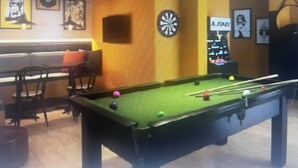 Biljardipöytä, seinässä tikanheittoalusta ja huoneessa myös jukebox, sohva ja tuoleja.