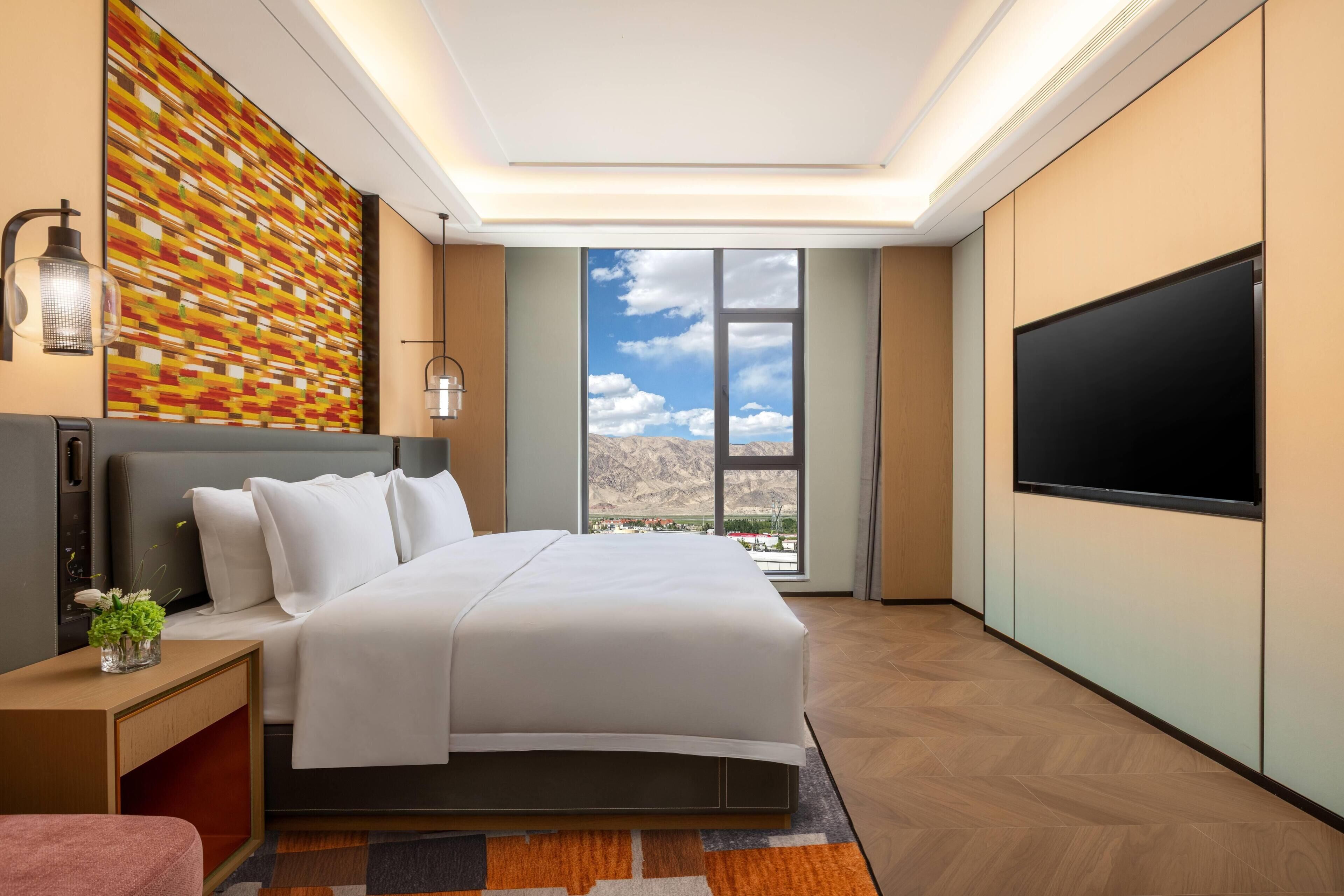 A modern hotel room with a large bed, a flat-screen TV, a view of mountains through the window, and a colorful wall art piece.