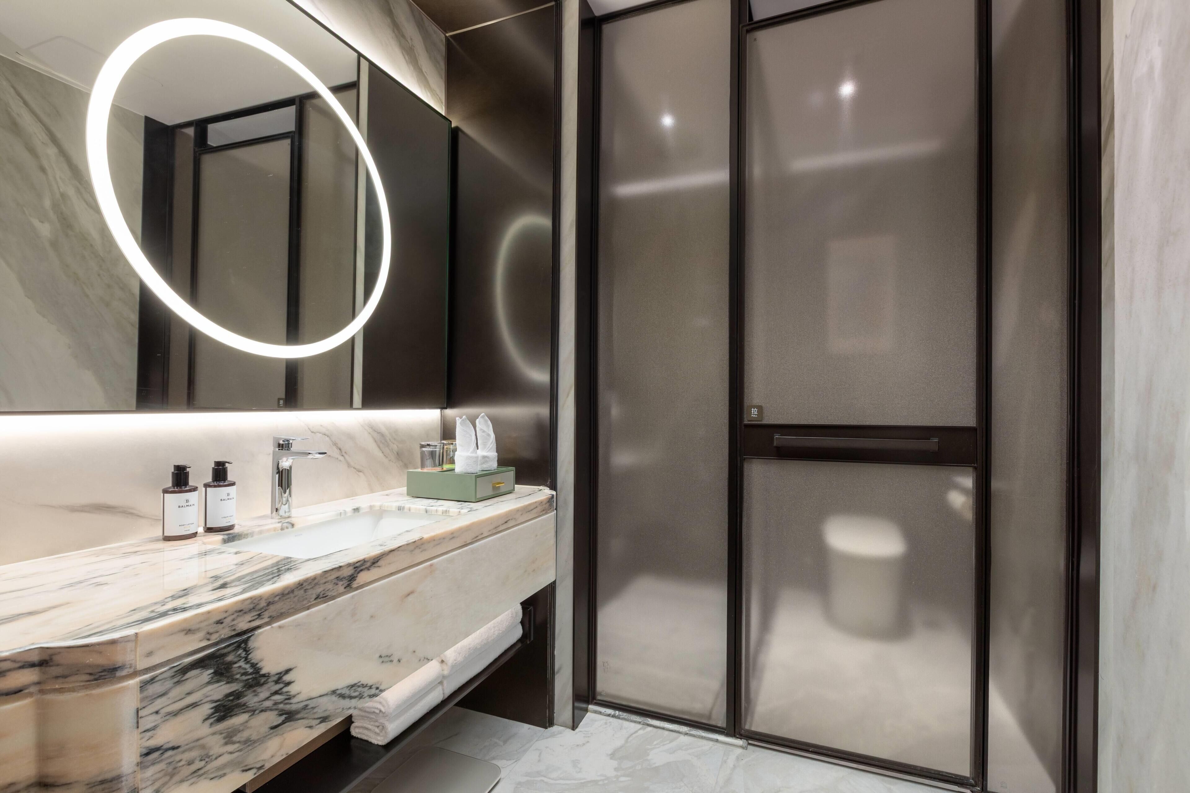 A modern bathroom with a marble countertop, a large mirror with a circular light, and a glass shower enclosure.