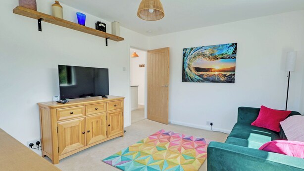 Een woonkamer met een televisie op een houten kast, een kleurrijk geometrisch vloerkleed, een groene bank met kussens, en een schilderij aan de muur.