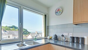 Een keuken met een raam, een spoelbak, houten kastjes, een waterkoker, een koffiezetapparaat en een klok aan de muur.