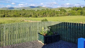 A view from a balcony with a wooden fence, a planter box with flowers, and a landscape of green fields and distant hills. - Spring Meadow Shepherds Hut (Rowrah)