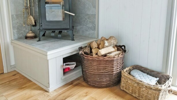 A room with a wood-burning stove, a basket of firewood, and a wicker basket with a cushion. - Spring Meadow Shepherds Hut (Rowrah)