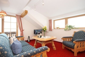 Ein Zimmer mit einer Couch, zwei Sesseln, einem Tisch mit Blumenvase, einem Fernseher und einem Fenster mit Vorhängen.