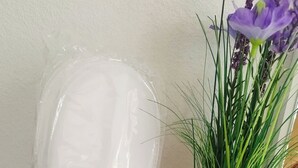 En fletkurv med en hvid etiket, der indeholder forskellige ting, heriblandt en lyserød æske og gule pensler. Ved siden af er en grøn vase med lilla blomster.