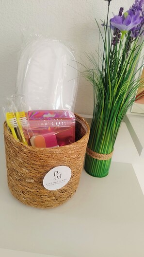 A wicker basket with a white label, containing various items including a pink box and yellow brushes. Next to it is a green vase with purple flowers.