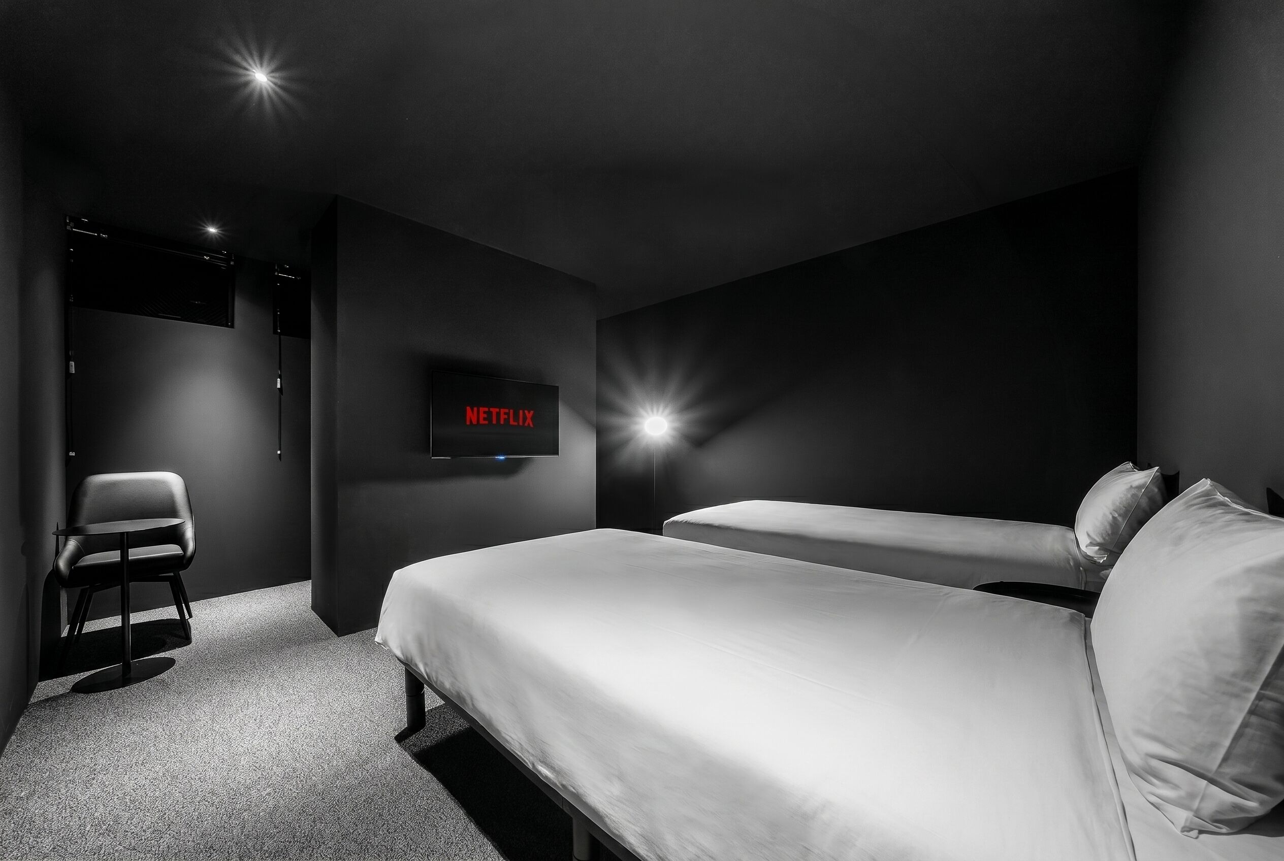 A modern hotel room with a single bed, a chair, a TV mounted on the wall, and a door leading to another room.