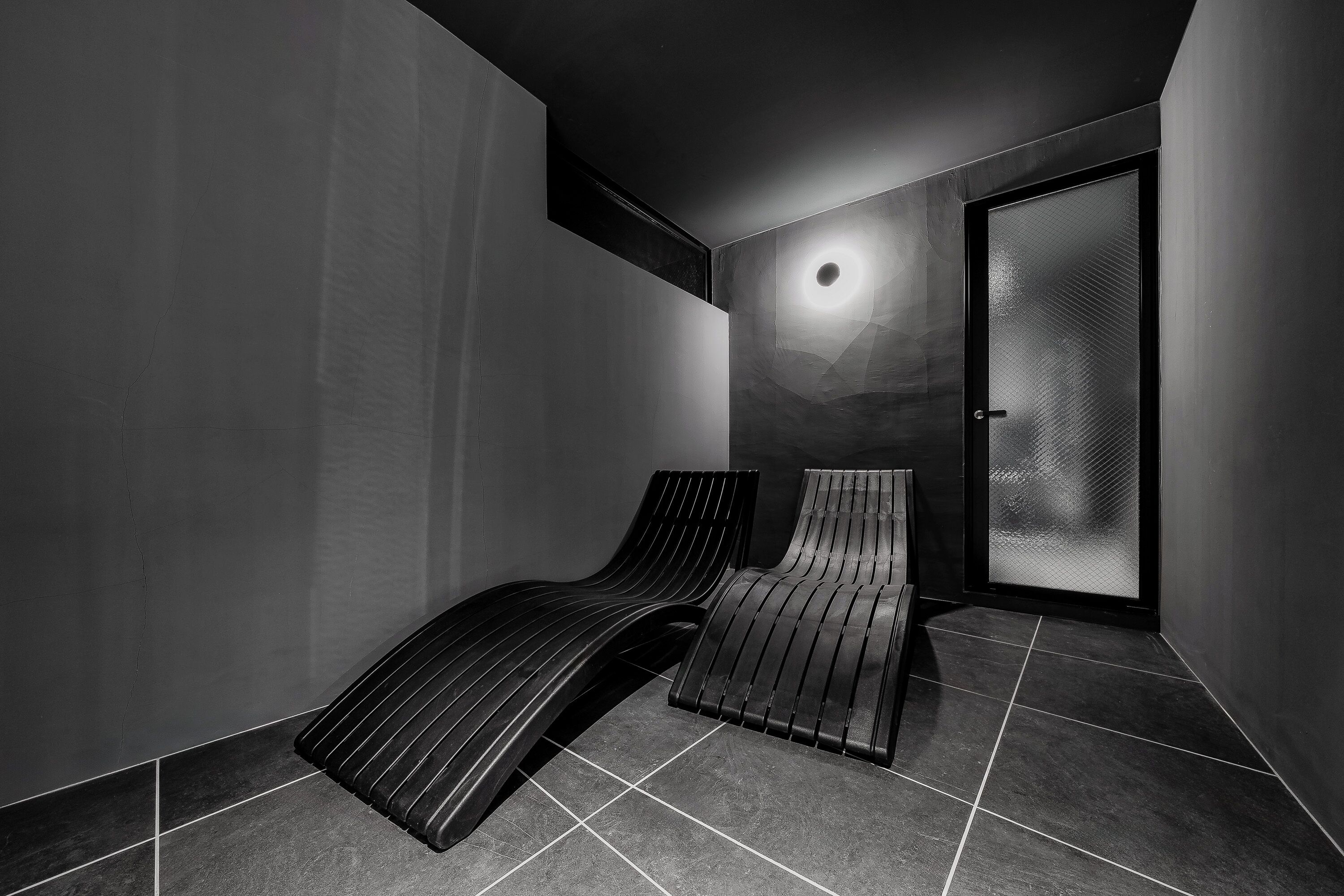 Una habitación moderna, con poca luz, una ventana grande, una puerta con vidrio esmerilado y dos sillones reclinables.