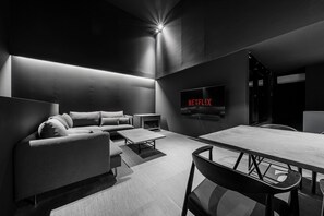 A modern, dimly lit room with a sofa, a coffee table, a dining table, and a flat-screen TV displaying 'NETFLIX'. - THE DWBL HOTEL B (Otaru)