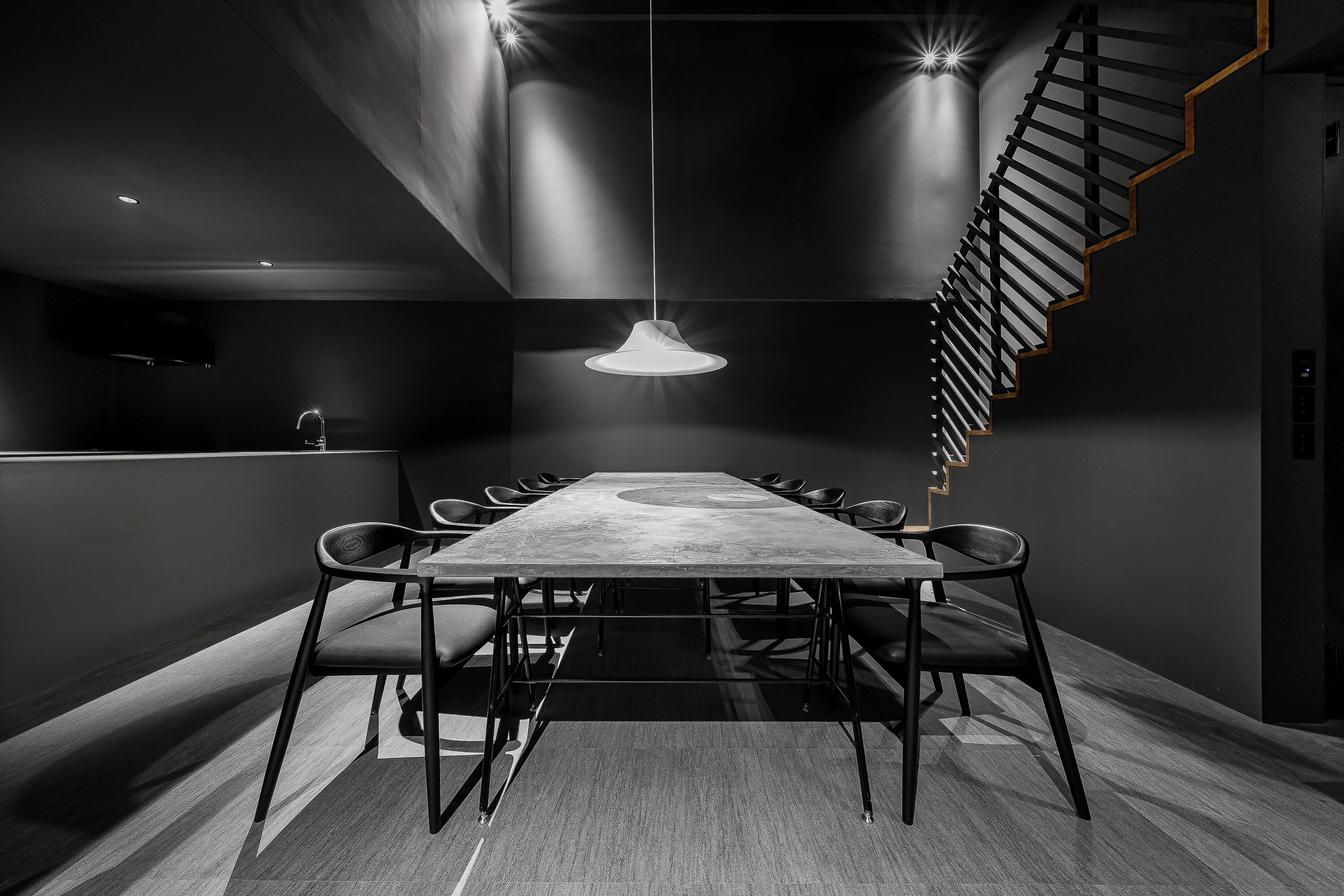 A modern dining area with a long table, chairs, a hanging light fixture, and a spiral staircase.