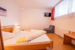 Ein Hotelzimmer mit zwei Betten, einem Schreibtisch, einem Sessel, einem an der Wand befestigten Fernseher und einem Fenster mit Vorhängen.
