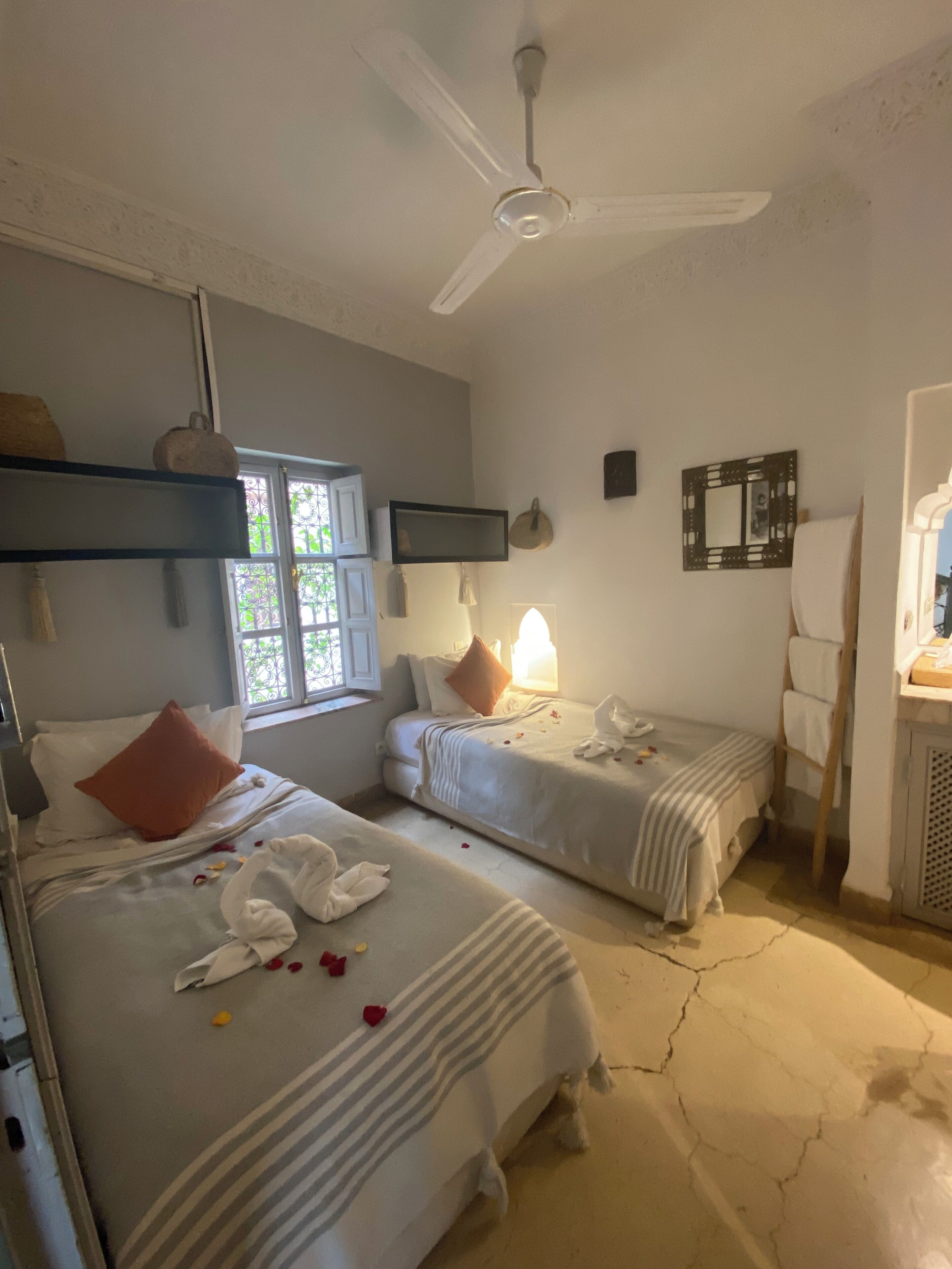 Hotellihuone, jossa on kaksi sänkyä, kattotuuletin, koristeellisella lasilla varustettu ikkuna ja pieni pöytä, jossa on lamppu.