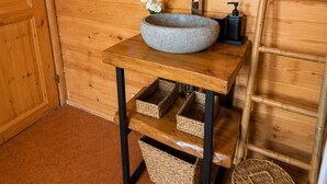 A wooden interior with a round mirror, a stone basin sink, wicker baskets, and a wooden ladder.