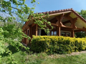 A wooden cabin with a covered porch, surrounded by lush greenery and a well-maintained hedge.