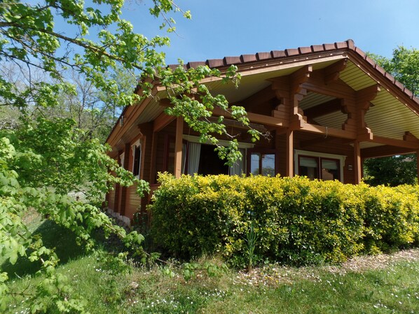 Une cabane en bois dotée d’un porche couvert, entourée d’une végétation luxuriante et d’une haie bien entretenue.