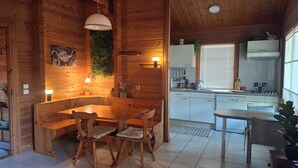 A wooden cabin interior with a dining table, chairs, a kitchen area, and a window with blinds.