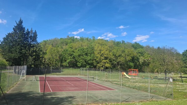 Un terrain de tennis avec un filet, entouré d’une clôture grillagée, et une aire de jeux avec un toboggan.