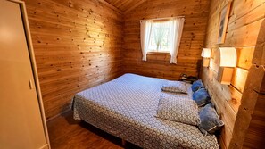 Une chambre dans un chalet en bois, avec un lit, des oreillers, une fenêtre avec des rideaux et une table de chevet avec une lampe.