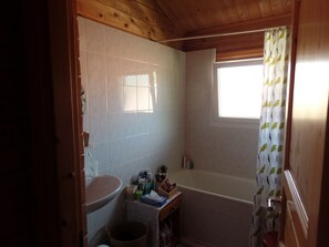 Une salle de bain équipée de toilettes, d’un lavabo et d’une baignoire. Il y a une fenêtre avec un rideau, et on peut voir des articles de toilette sur le lavabo.