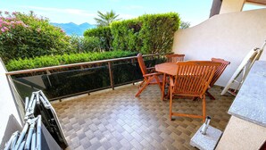 A balcony with a wooden table and chairs, a glass balustrade, and a view of greenery and mountains. - Lake View Apartment in Luino for 4 (Luino)