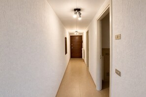 Ein schmaler Gang mit einer Holztür am Ende, einer Lampe darüber und einem Lichtschalter auf der rechten Seite.
