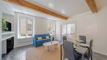 A modern living room with a blue sofa, a dining table set for two, and a TV on a shelf.