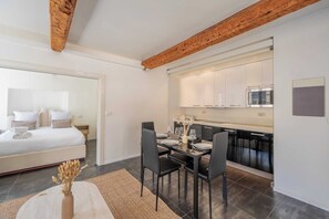 A modern bedroom with a bed, bedside table, and a nightstand. Adjacent is a dining area with a table, chairs, and a vase of dried flowers. The room features exposed wooden beams and a large window. - 1BDR/4P Apt in the Heart of Saint-Tropez-near Port (Saint-Tropez)