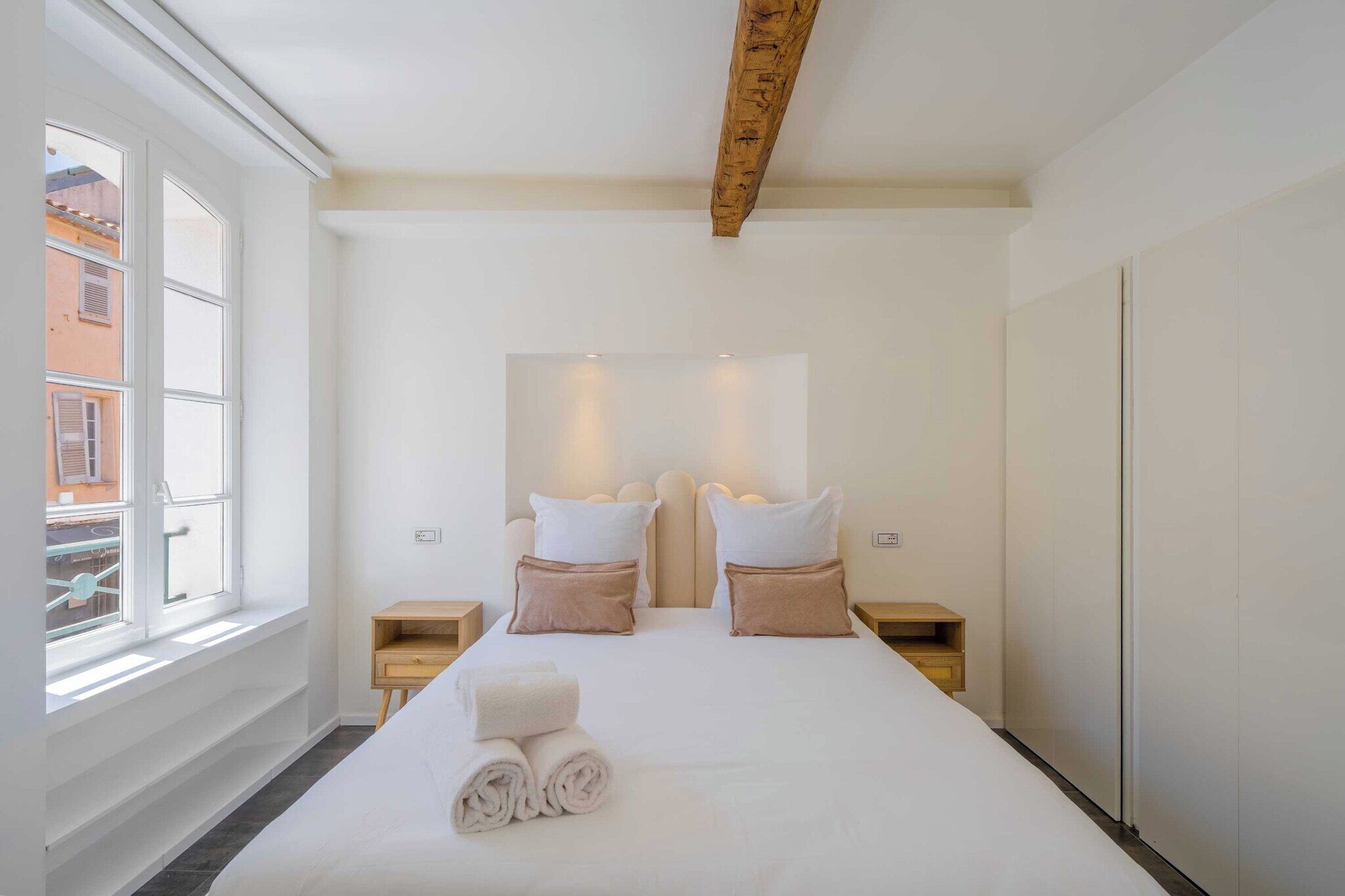 A hotel room with a large bed, two bedside tables, a wooden ceiling beam, and a window with a view of buildings.