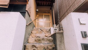 A stone staircase leading to a wooden door with a traditional design, a small outdoor area with a stone bench, and a wall-mounted light fixture. - Shinobi House – 4-Bedroom Ninja-Inspired Stay (大阪市)