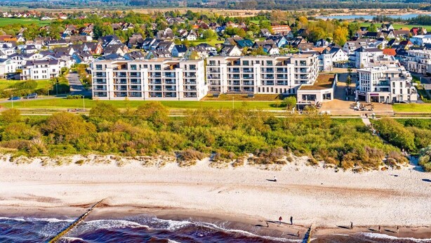 Ett bostadsområde vid kusten med moderna lägenhetsbyggnader, en sandstrand och fri utsikt över havet.