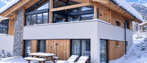 Une maison moderne en bois à deux étages, dotée de grandes fenêtres et d’un balcon, entourée de neige.