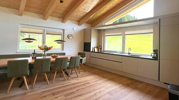 Ett modernt kök med träbjälkar, en matplats med stolar, ett fönster med utsikt över grönområden och en klocka på väggen.