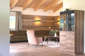 A modern interior with a wooden ceiling, a sofa, a pink chair, a small table with a vase, and a cabinet with glass doors. - Holiday home directly on the ski slope with sauna (St. Lambrecht)