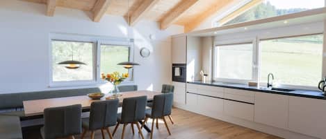 A modern kitchen with wooden beams, a dining area with a table and chairs, and large windows offering a view of the outdoors.