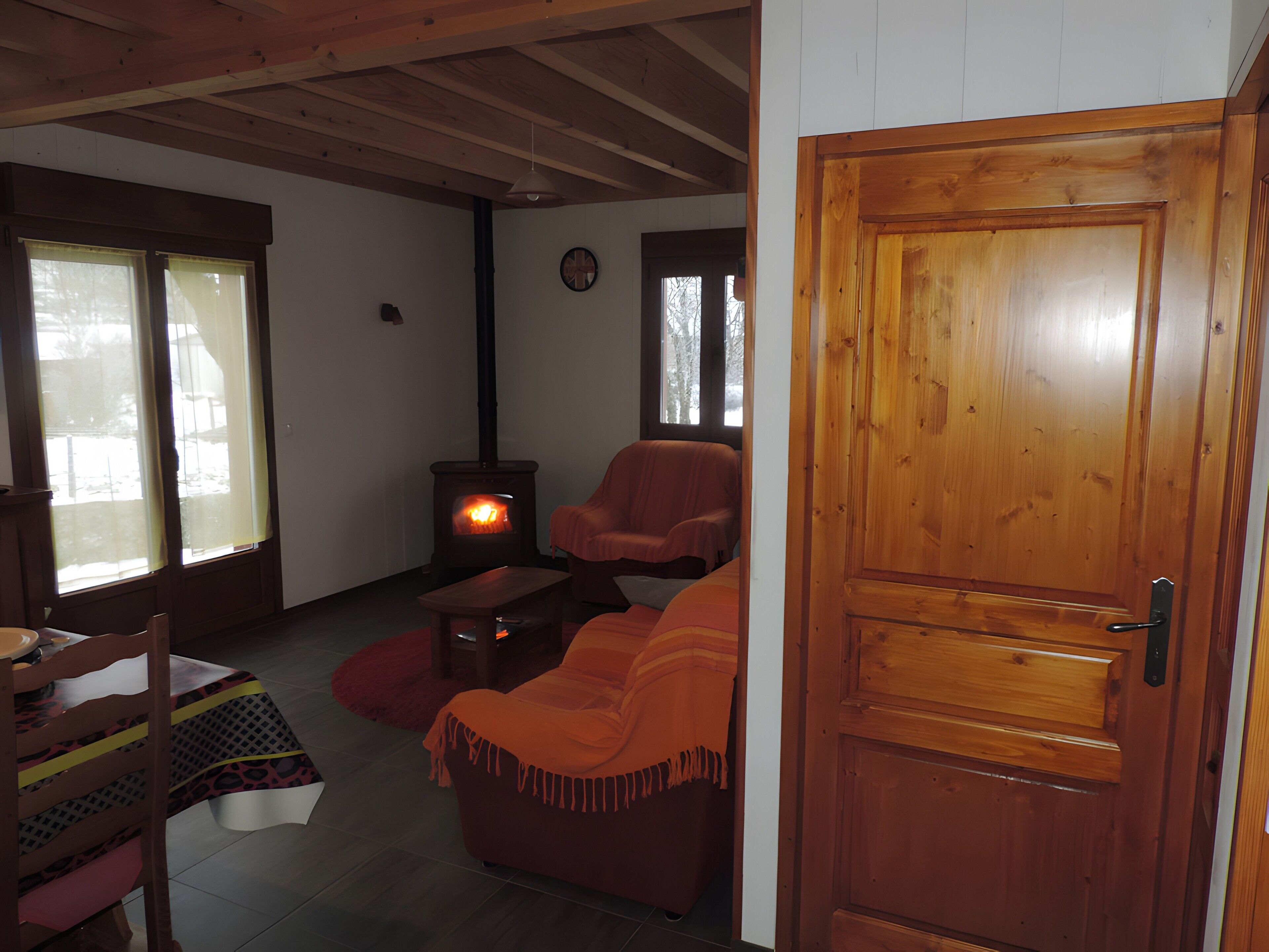 A cozy living room with a wooden ceiling, a sofa, a chair, a small table, and a wood-burning stove.