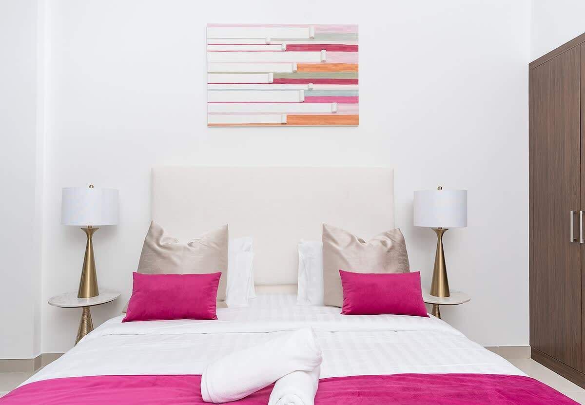 A neatly made bed with white and pink bedding, two bedside lamps, and a wooden wardrobe.