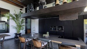 A modern kitchen with dark cabinetry, a central island, and a seating area with wicker chairs. - Villa Suwan -The Hilltop Escape Samui (Koh Samui)