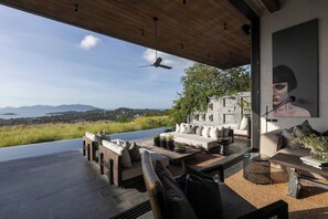 A modern outdoor living space with a view of the sea, a ceiling fan, and a large painting. - Villa Suwan -The Hilltop Escape Samui (Koh Samui)