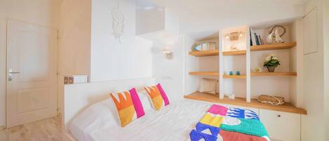 A bedroom with a bed, pillows, a bedside table, a shelf with books and decorative items, and a wall-mounted light.