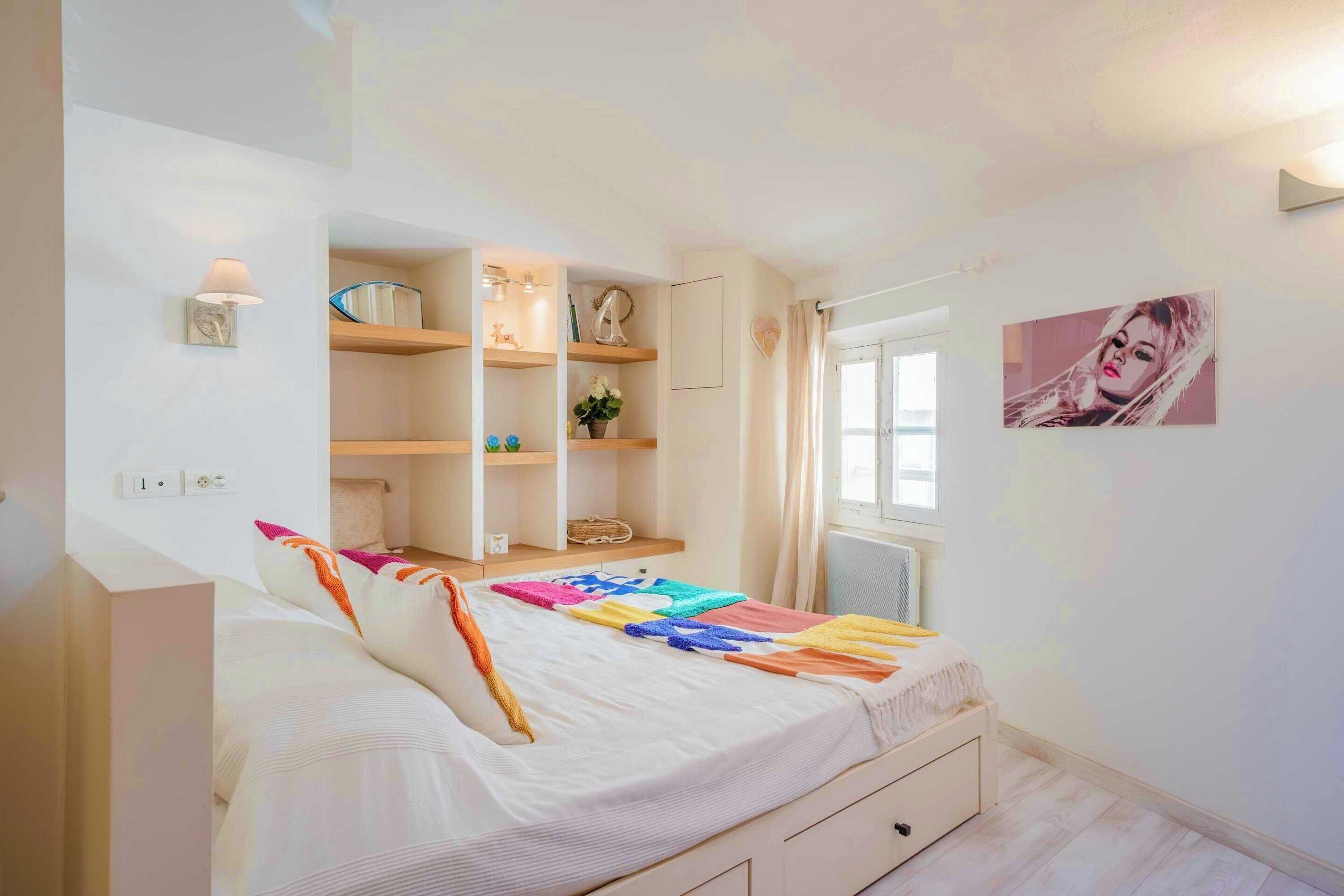 Een slaapkamer met een bed, een inbouwkleerkast, een nachtkastje, een raam met gordijnen en een wanddecoratie.