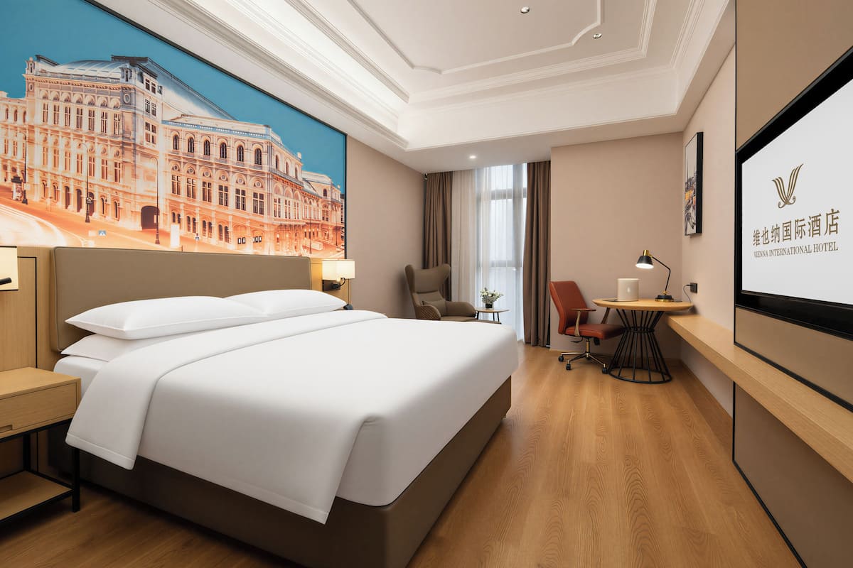 A hotel room with a large bed, a desk, a chair, and a view of a building through the window.
