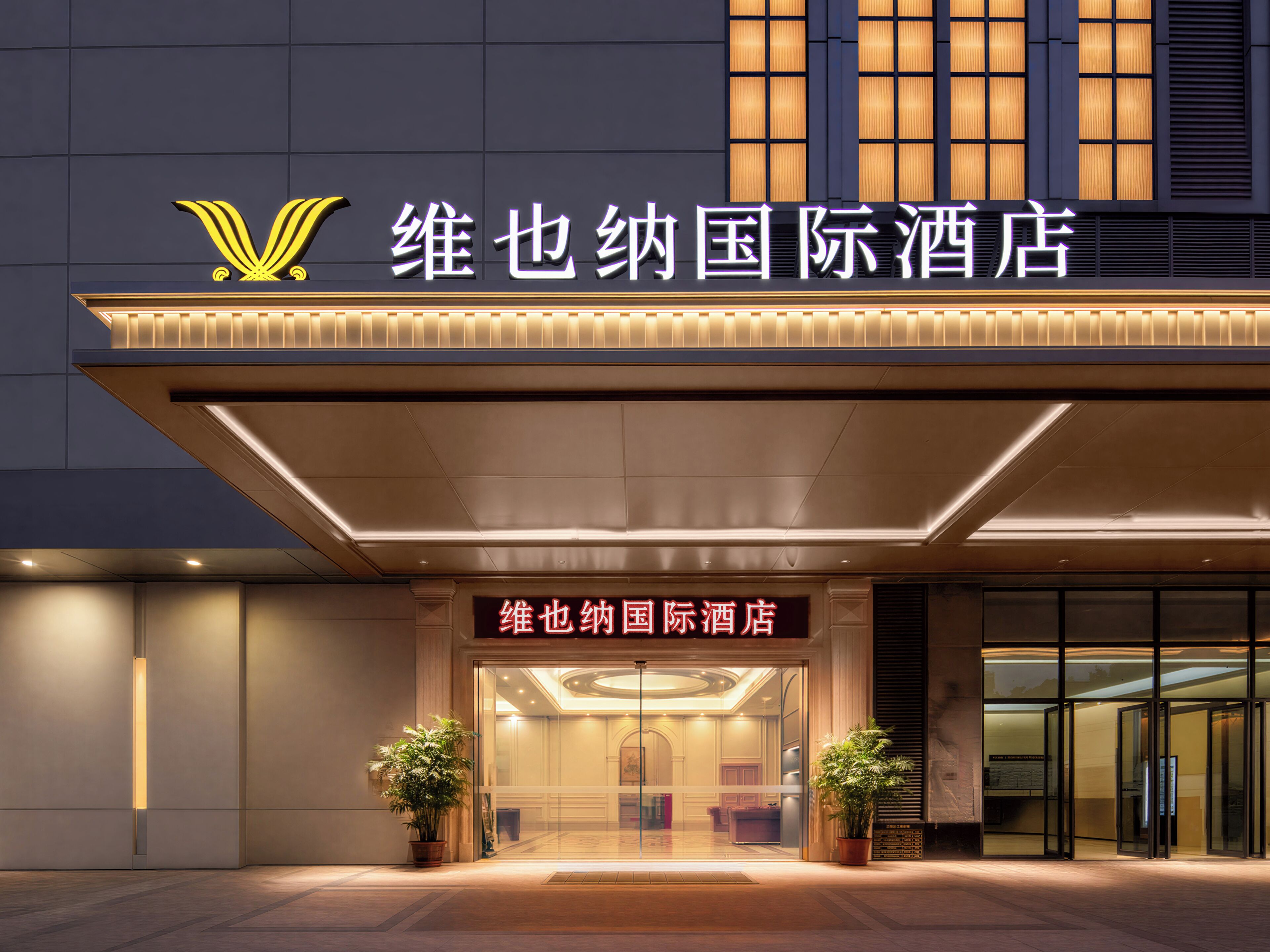 A hotel entrance with a sign, a logo, and two potted plants.