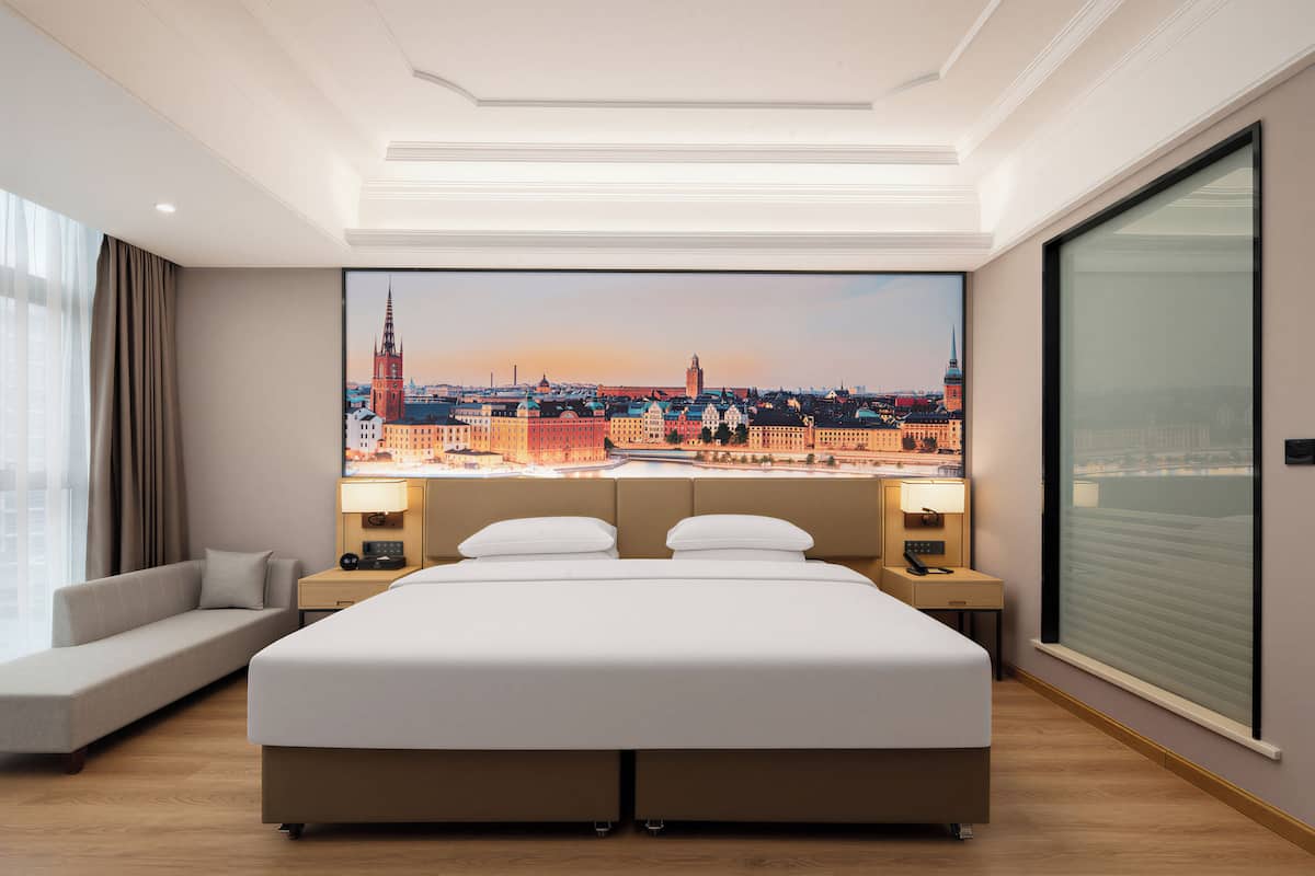 A hotel room with a large bed, a sofa, a cityscape view, and a large window.