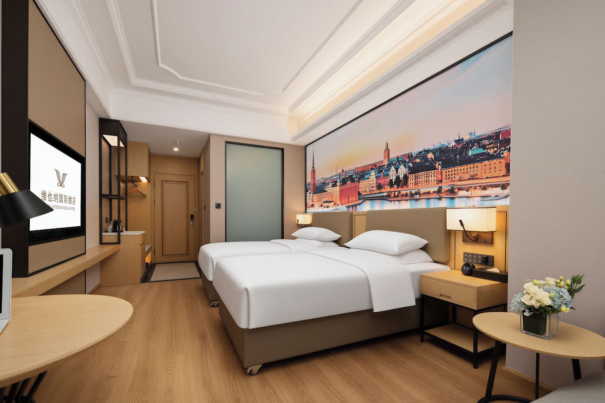 A hotel room with a bed, bedside table, lamp, and a painting of a cityscape on the wall.