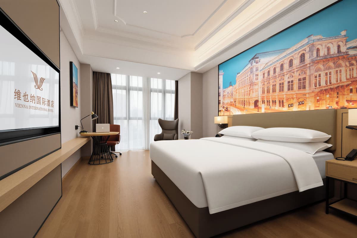 A hotel room with a large bed, a desk, a chair, a TV screen, and a large window.