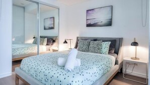 Un dormitorio con cama, mesita de noche, lámpara, espejo y un cuadro en la pared.